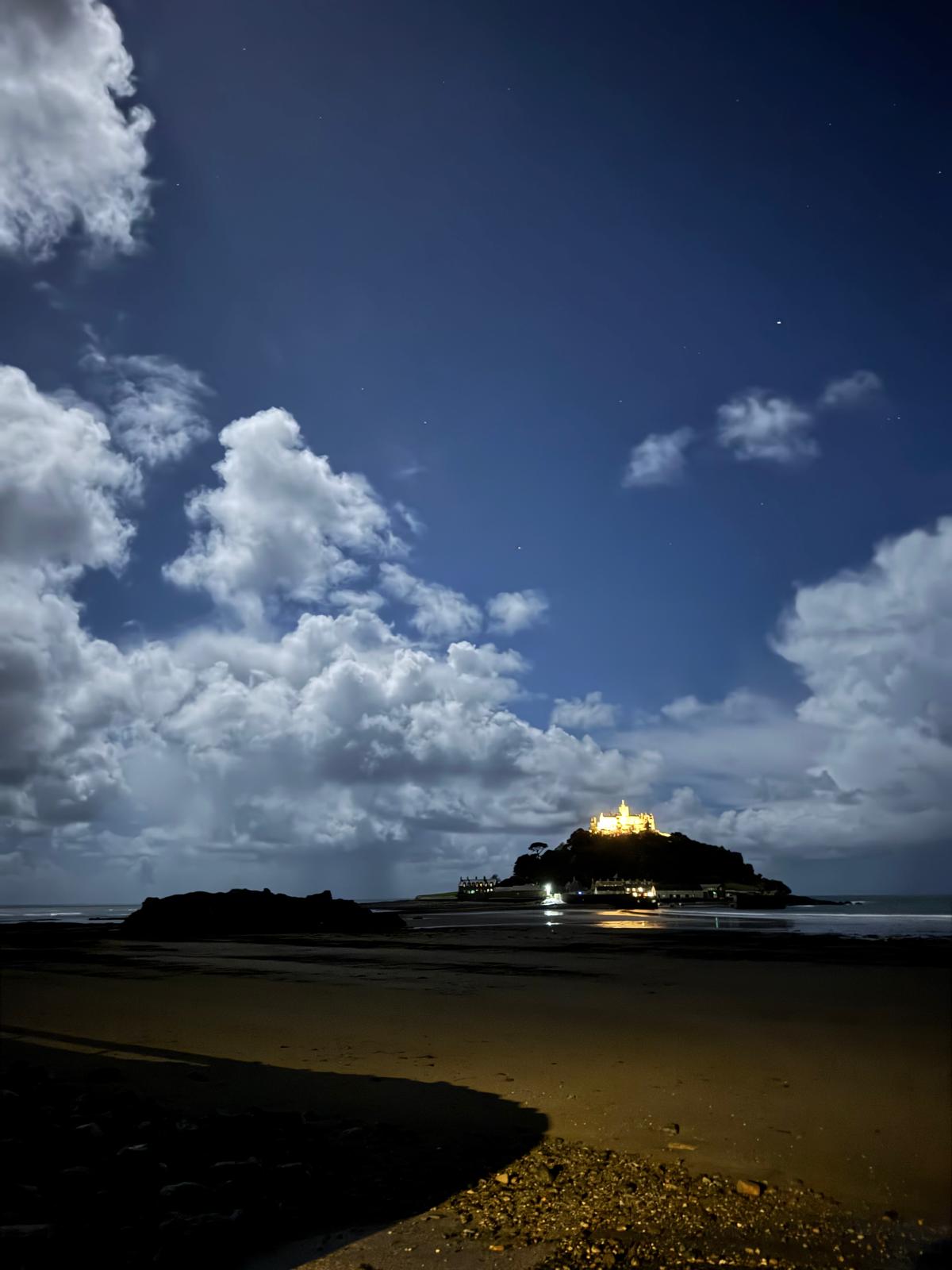 St Michael’s Mount illuminated at night beneath clouds, with light reflected on the wet sand